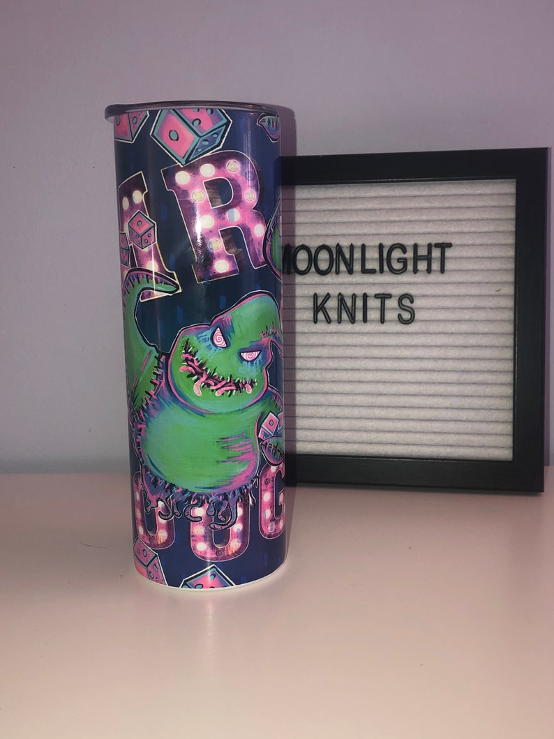 Puede incluir: Un vaso azul y verde con un gr&aacute;fico de un personaje de la pel&iacute;cula "Pesadilla antes de Navidad". El personaje est&aacute; rodeado de luces rosas y blancas que deletrean "Boo".