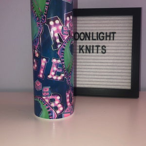 Puede incluir: Un vaso de acero inoxidable azul y verde con un dise&ntilde;o gr&aacute;fico rosa y blanco de la palabra "MAGIC" en luces de ne&oacute;n y dados.