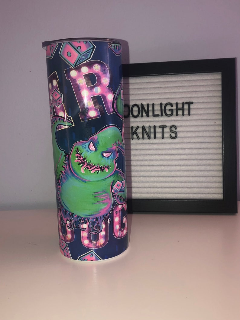 Puede incluir: Un vaso azul y verde con un gr&aacute;fico de un personaje de la pel&iacute;cula "Pesadilla antes de Navidad". El personaje es verde con costuras rosas y sostiene un dado. El vaso tiene la palabra "Boo" escrita en letras rosas con luces blancas.