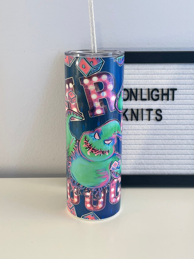 Puede incluir: Un vaso de acero inoxidable azul y verde con un gr&aacute;fico de un monstruo verde con la boca cosida y las palabras "Oogie Boogie" en luces rosas y blancas. El vaso tiene una tapa transparente y una pajita plateada.