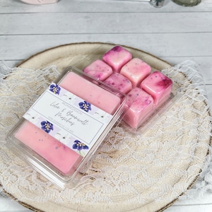 Könnte beinhalten: Zwei transparente Kunststoffbehälter mit rosa Wachsschmelzen. Ein Behälter enthält einen rechteckigen Riegel, der andere rautenförmige Würfel. Auf dem Etikett steht "Lily & Baumwoll Moschus". Die Schmelzen liegen auf einer Holzscheibe.