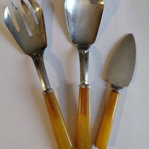 May include: A vintage salad serving set featuring a fork, spoon, and pie server. The utensils have silver-toned heads and amber-colored handles. The fork has three tines, and the spoon has a wide, flat bowl.