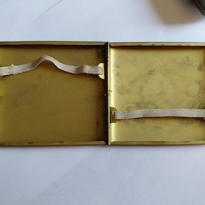 May include: Open, vintage brass cigarette case. The interior is a golden color with two elastic bands. The case is rectangular with a hinged opening and a clasp. The case is empty.