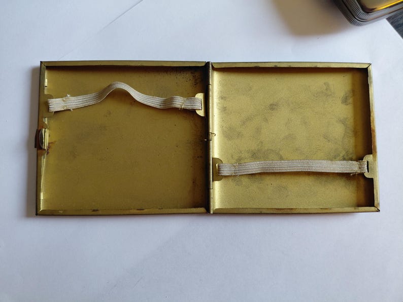 May include: Open, vintage brass cigarette case. The interior is a golden color with two elastic bands. The case is rectangular with a hinged opening and a clasp. The case is empty.