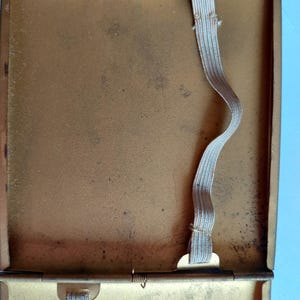 May include: Open, vintage gold-toned metal cigarette case with a hinged lid. The interior features two elastic bands for securing cigarettes. The case shows signs of age with some discoloration and wear, suggesting it is a used item.