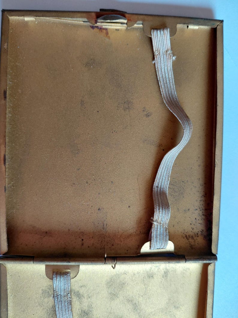 May include: Open, vintage gold-toned metal cigarette case with a hinged lid. The interior features two elastic bands for securing cigarettes. The case shows signs of age with some discoloration and wear, suggesting it is a used item.