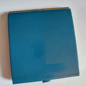 May include: A vintage teal compact mirror with a rectangular shape. The compact has a gold-colored hinge and a small, circular clasp. The surface of the compact shows some wear and tear, adding to its antique charm.