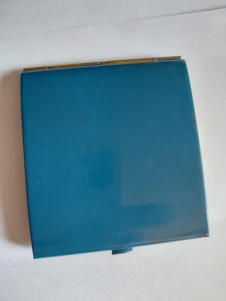 May include: A vintage teal compact mirror with a rectangular shape. The compact has a gold-colored hinge and a small, circular clasp. The surface of the compact shows some wear and tear, adding to its antique charm.