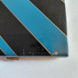 May include: A rectangular, vintage metal box with a black and light blue diagonal stripe pattern. The box has a gold-colored clasp and shows signs of wear, with some paint chipping on the edges.