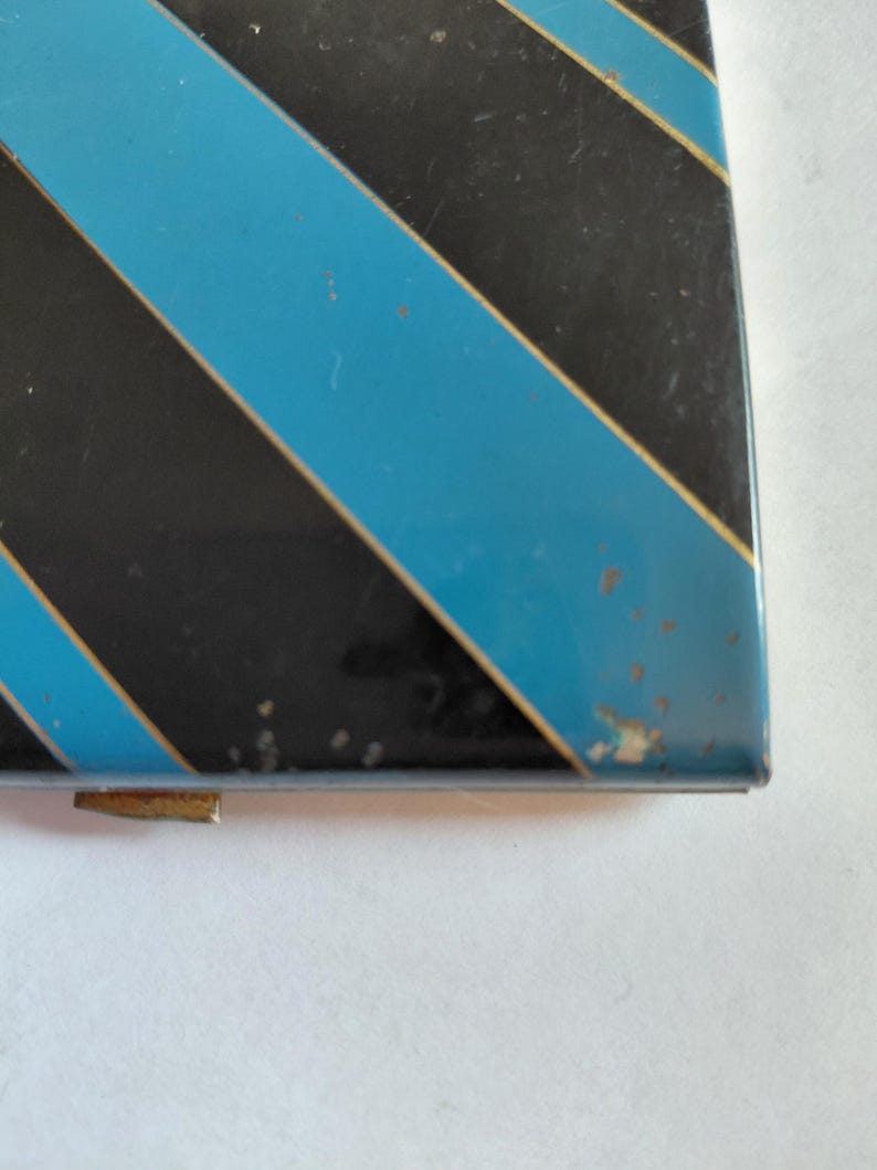 May include: A rectangular, vintage metal box with a black and light blue diagonal stripe pattern. The box has a gold-colored clasp and shows signs of wear, with some paint chipping on the edges.