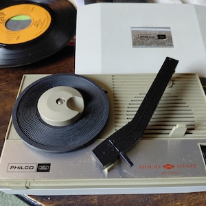 May include: A vintage Philco Ford solid state 45 RPM record player featuring a beige plastic casing, a black circular turntable platter, and a black tonearm. The control panel includes the Philco Ford logo and the text Solid State 45 RPM.