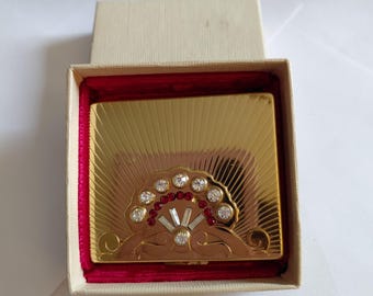 May include: A gold-toned compact mirror with a sunburst design, adorned with clear and red rhinestones in a fan-shaped pattern. The compact is presented in a cream-colored box with a red velvet lining, suggesting a vintage or collectible item.