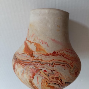 May include: A small, rounded ceramic vase with a matte, light beige upper section and a wide, bulbous base. The lower portion features a swirling, marbled pattern of vibrant orange, deep red, and dark brown paint, creating a fluid, layered effect.
