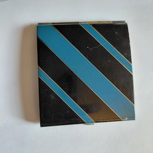 May include: A vintage compact mirror with a black and teal diagonal stripe design. The compact has a rectangular shape with a gold trim. The design is a bold, geometric pattern. The compact is closed and appears to be in good condition.