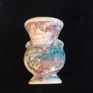 May include: A small, decorative vase with a unique shape and marbled paint effect. The vase features a white base with swirls of red, blue, and purple. It has two small handles on the sides and a flared top and base.