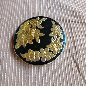 May include: A round, black compact with a gold floral design. The flowers are embossed and have a raised texture.
