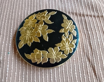 May include: A round, black compact with a gold floral design. The flowers are embossed and have a raised texture.