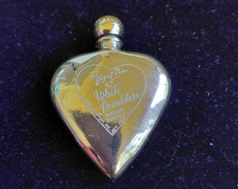 May include: A heart-shaped, silver-toned perfume bottle with a rounded cap. The bottle features the text "Byan White Shoulders Perfume Essence" in a heart-shaped design. The bottle is approximately 3 inches long.