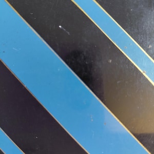 May include: Black surface with diagonal stripes in alternating colors of blue and black, with thin gold stripes bordering the blue. The design is a repeating pattern.