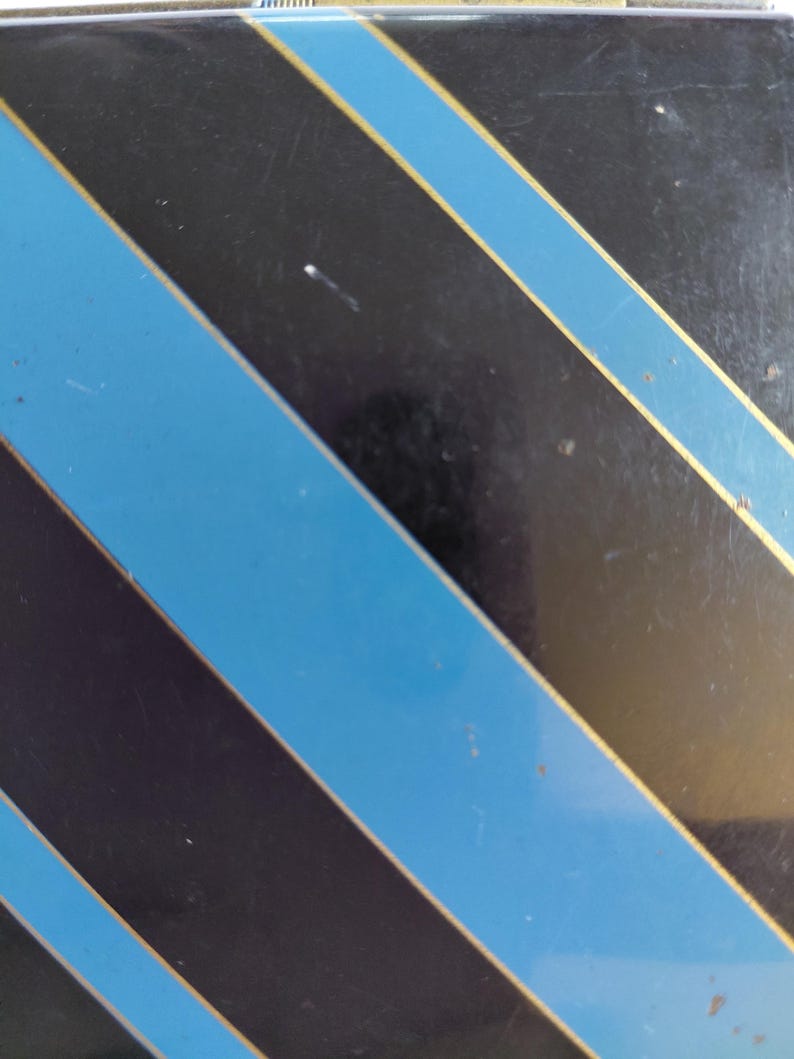 May include: Black surface with diagonal stripes in alternating colors of blue and black, with thin gold stripes bordering the blue. The design is a repeating pattern.