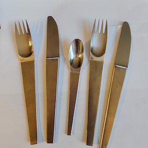 May include: A set of five stainless steel utensils: two forks, two knives, and a spoon. The utensils have long, flat handles and a simple, modern design. The knives have serrated blades. The spoon has a rounded bowl.
