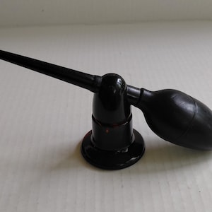 May include: A black rubber bulb duster with a long, slender nozzle attached to a circular base. The device features a flexible, pear-shaped squeeze bulb connected to a rigid, pointed tip, designed for precision air application in a compact form.