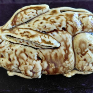 May include: A ceramic rabbit figurine in a lying down position. The rabbit is primarily cream-colored with brown mottled markings. The figurine has detailed features, including ears and a small tail. The overall design is smooth and glossy.