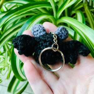 May include: A black crochet bat keychain with gray ears and wings. The bat has a silver key ring attached. The keychain is held in a hand, with green and white foliage in the background.