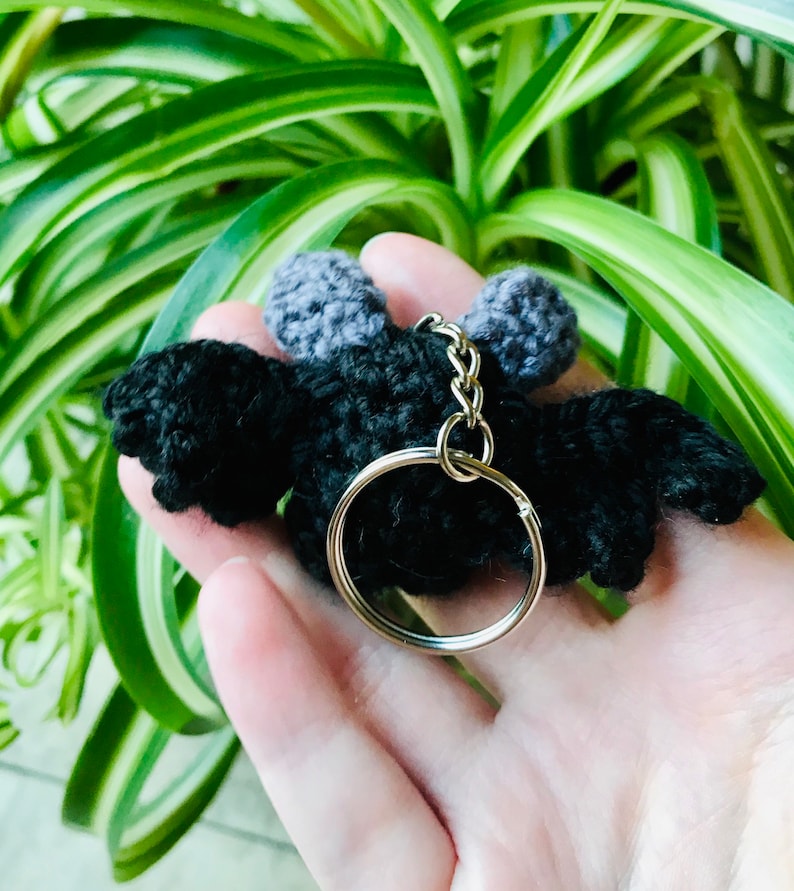May include: A black crochet bat keychain with gray ears and wings. The bat has a silver key ring attached. The keychain is held in a hand, with green and white foliage in the background.