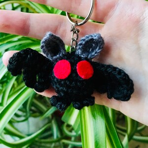 May include: A handmade black crochet moth keychain with red eyes and gray antennae. The keychain is attached to a silver metal ring. The moth has a fluffy texture and is held against a background of green leaves.