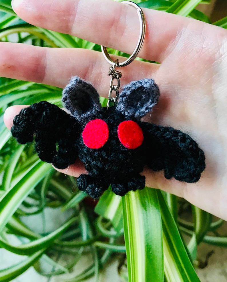 May include: A handmade black crochet moth keychain with red eyes and gray antennae. The keychain is attached to a silver metal ring. The moth has a fluffy texture and is held against a background of green leaves.