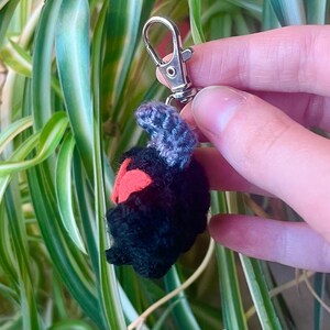 May include: A handmade crochet keychain featuring a black creature with red accents and a gray top. The keychain is attached to a silver-colored metal clasp. The background includes green and white striped leaves.