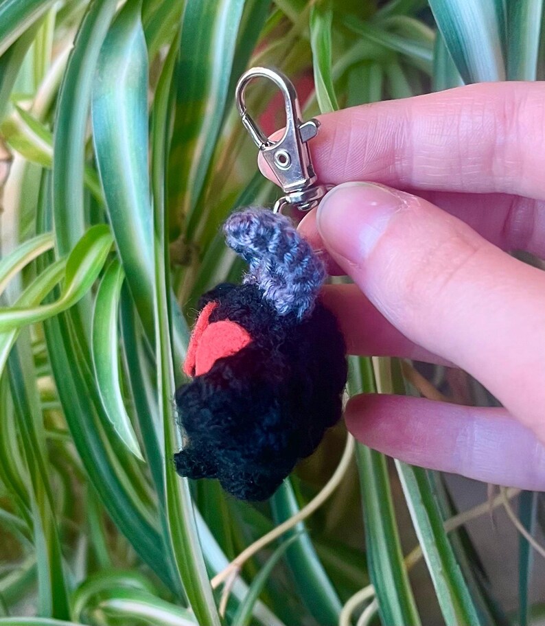 May include: A handmade crochet keychain featuring a black creature with red accents and a gray top. The keychain is attached to a silver-colored metal clasp. The background includes green and white striped leaves.