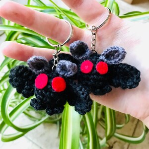 May include: Two handmade crochet moth keychains. The moths are black with gray wings and red eyes. Each keychain has a silver metal ring and chain. The keychains are held up against a background of green and white leaves.