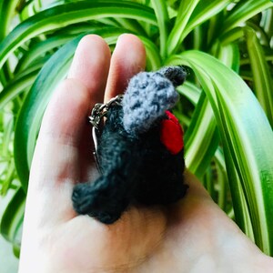 May include: A handmade black crochet keychain shaped like a bat, with gray wings and a red heart. The keychain is held in a hand, with a green and white leafy background.