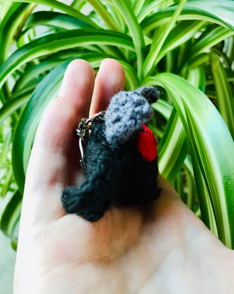 May include: A handmade black crochet keychain shaped like a bat, with gray wings and a red heart. The keychain is held in a hand, with a green and white leafy background.