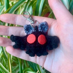 May include: A handmade black crochet keychain featuring a moth-like creature with red eyes and gray antennae. The keychain has a silver clasp for easy attachment to keys or bags. The moth has a unique, whimsical design.