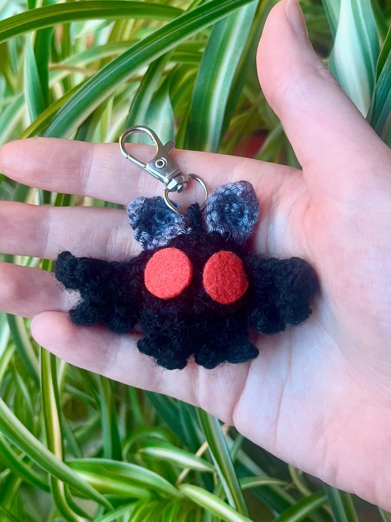May include: A handmade black crochet keychain featuring a moth-like creature with red eyes and gray antennae. The keychain has a silver clasp for easy attachment to keys or bags. The moth has a unique, whimsical design.