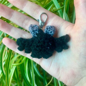 May include: A handmade crochet bat keychain in black with gray accents. The bat has outstretched wings and a silver-colored metal clasp. The keychain is held in a hand, with green and white foliage in the background.