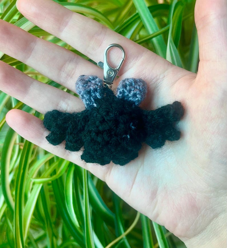 May include: A handmade crochet bat keychain in black with gray accents. The bat has outstretched wings and a silver-colored metal clasp. The keychain is held in a hand, with green and white foliage in the background.