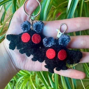 May include: Two handmade black crochet keychains shaped like a moth, each with two red circular eyes and gray antennae. The keychains are attached to silver-colored metal rings and clasps. The background features green and white foliage.