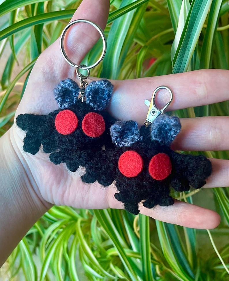 May include: Two handmade black crochet keychains shaped like a moth, each with two red circular eyes and gray antennae. The keychains are attached to silver-colored metal rings and clasps. The background features green and white foliage.