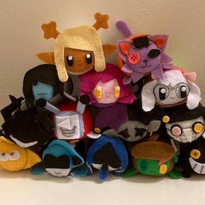 May include: A collection of handmade felt plushies in various colors, featuring characters with distinct facial expressions and designs. The plushies include characters with yellow, blue, black, and purple features, stacked together.