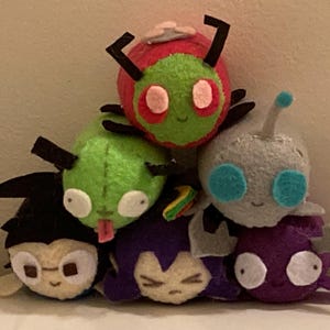 May include: A pile of handmade plush toys in various colours. The toys are stacked, featuring characters with large eyes and antennae. The plushies are primarily green, red, purple, and grey.