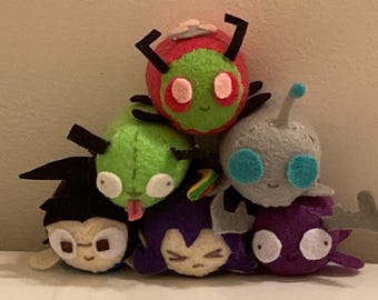 May include: A collection of handmade plush toys in various colors. The toys are stacked on top of each other, featuring characters with large eyes and antennae. The plushies are primarily green, red, purple, and gray.