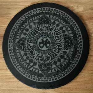 Könnte beinhalten: Ein runder, schwarzer Schieferuntersetzer mit einem geätzten Mandala-Design. Das komplizierte Muster weist florale und geometrische Elemente mit einem zentralen Marienkäfermotiv auf. Der Untersetzer steht auf einer Holzoberfläche.