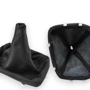 May include: Two black leather car shift boots. One is upright, showing the cone shape and stitching. The other is open, revealing the interior and white clips. Automotive parts.
