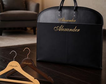 May include: A black garment bag with two top handles and the name Alexander embroidered in gold script. Two wooden clothes hangers are placed in front, one light wood and one dark wood, both featuring the name Alexander in gold.