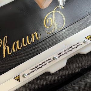 May include: An embroidery machine needle stitching the yellow cursive name Shaun D onto black faux leather. A white plastic embroidery hoop with magnetic warning labels reading Keep Away From Electronic Devices, and Credit Cards.