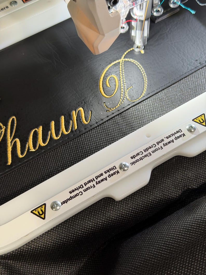 May include: An embroidery machine needle stitching the yellow cursive name Shaun D onto black faux leather. A white plastic embroidery hoop with magnetic warning labels reading Keep Away From Electronic Devices, and Credit Cards.